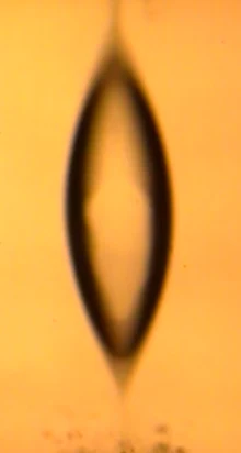 Drop stretched into a spindle with pointed ends, shedding particles from its lower tip.