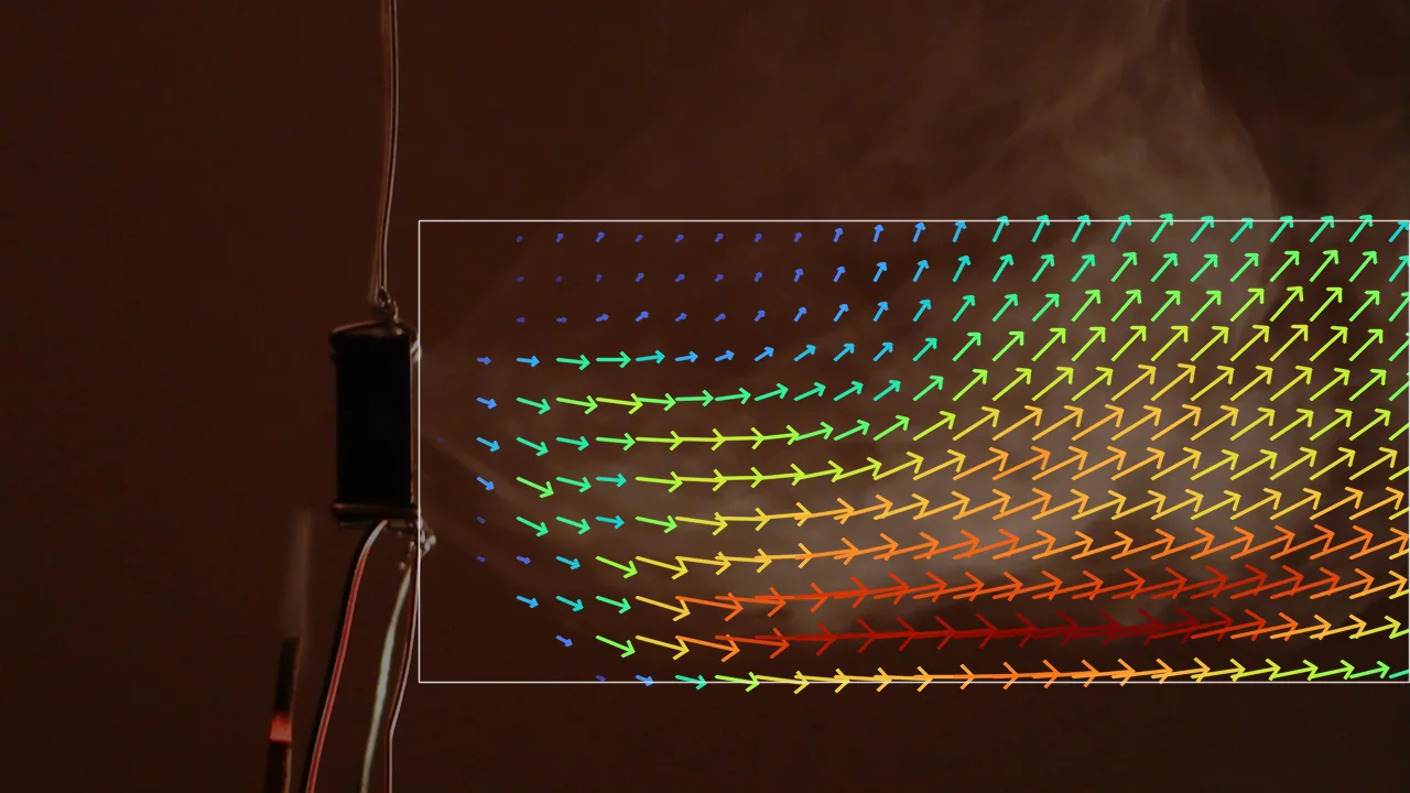 The test frame, dimmed, with arrows of the 40-second mean velocity in the region right of the fan: short blue arrows near the outlet, and longer green to red arrows downstream pointing right, turning up and to the right in the upper part.